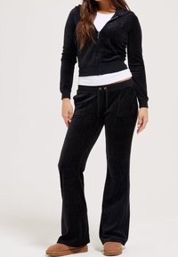Black velour tracksuit with zip-up hoodie and drawstring pants. Slim fit, soft texture. Model wearing tan slippers.