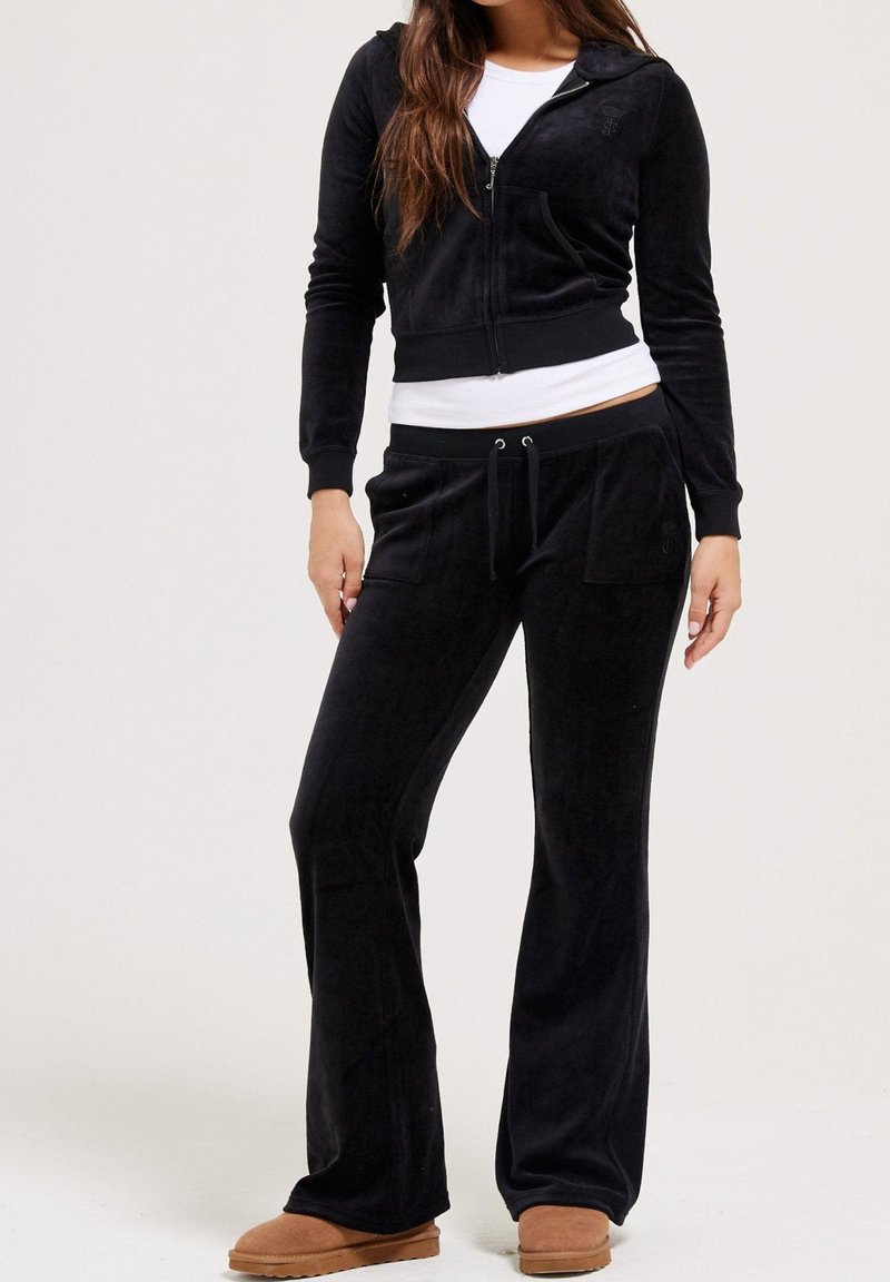 Black velour tracksuit with zip-up hoodie and drawstring pants. Slim fit, soft texture. Model wearing tan slippers.