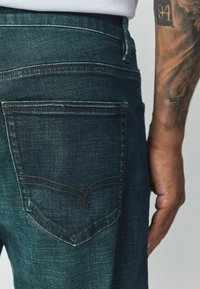 Tume sinised teksapüksid, millel on tagataskul eristuva kumerjoonelise õmblemisega disain. Tekstuur tundub sile ja kerge pleekimisega.