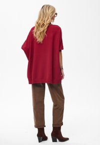 Czerwony, dziany, oversize sweter z krótkimi rękawami, zestawiony z brązowymi spodniami z podwiniętymi mankietami oraz burgundowymi botkami na kostkę z blokowym obcasem.