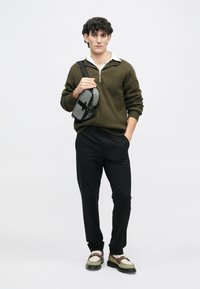 Olive green knitted sweater, white collared shirt, black trousers, and two-tone shoes. Holding a gray patterned bag with a black strap.