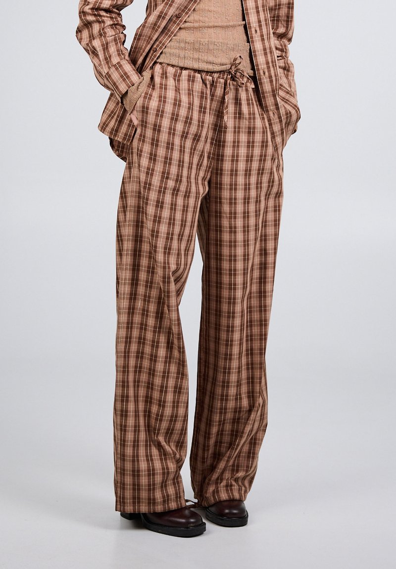 Barna kockás, széles szárú nadrág húzózsinóros derékrésszel, hozzáillő kockás inggel, bézs pulóverrel és sötétbarna cipővel, semleges háttér előtt.