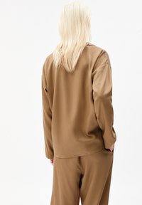 Persona con lunghi capelli biondi che indossa una maglietta a maniche lunghe marrone larga e pantaloni abbinati, voltata di spalle su uno sfondo bianco.