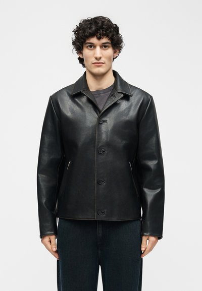 Άνδρας με σγουρά μαλλιά, φοράει μαύρο δερμάτινο μπουφάν με κουμπιά και σκούρο παντελόνι, στέκεται μπροστά σε απλό ανοιχτό γκρι φόντο.