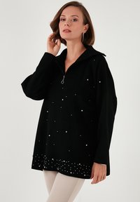 Musta neule, jossa on puolivetoketju kauluksessa, savuinen hopeanväristen koristeiden kanssa ja koristeellinen alaosa, jossa on helmimäinen kuvio.