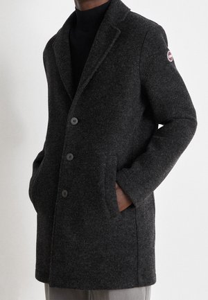 Homme portant un manteau en laine gris foncé boutonné, les mains dans les poches et un écusson logo sur la manche gauche supérieure.