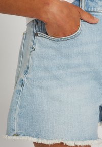 Pantalones cortos de mezclilla azul claro con dobladillo deshilachado, diseño de cinco bolsillos y corte ajustado. Costuras de los bolsillos visibles y detalles de herrajes metálicos.