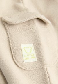 Beige szövet, finom kötött textúrával, amelyen egy fedeles zseb található. Egy címkén egy medve design és a "// LITTLE ONE - 365" felirat látható világos zöld színben.