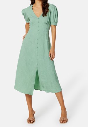 Femme portant une robe midi vert clair texturée avec boutons sur le devant, manches ballon et encolure en V, associée à des sandales beiges tissées à talons.
