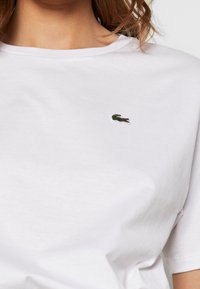 Weißes Baumwoll-T-Shirt mit rundem Ausschnitt, das auf der linken Brustseite ein kleines, grünes gesticktes Krokodil-Logo zeigt. Kurze Ärmel.
