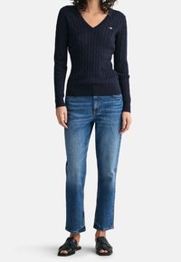 Vrouw in een marineblauwe kabelgebreide V-halstrui, blauwe recht-toe-jeans en zwarte geweven sandalen met open teen staat tegen een witte achtergrond.