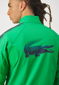 Zöld dzseki texturált anyagból, hálós részletekkel, magas gallérral és fekete krokodil logóval, amelynek geometrikus mintázata van a hátsó részen.