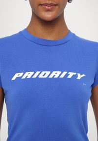 T-shirt blu a maniche corte in cotone, con la scritta bianca in grassetto "PRIORITY" sul petto, dalla superficie liscia e collo rotondo.