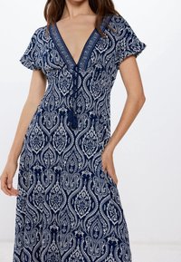 Robe bleu marine avec un motif cachemire blanc, col en V, manches courtes volantées et taille cintrée. Comprend un détail à pompons sur un lien.