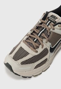 Baskets Nike Vomero 5 en maille beige et marron avec accents noirs. Comprend des superpositions texturées, des lacets plats et une semelle en caoutchouc durable.