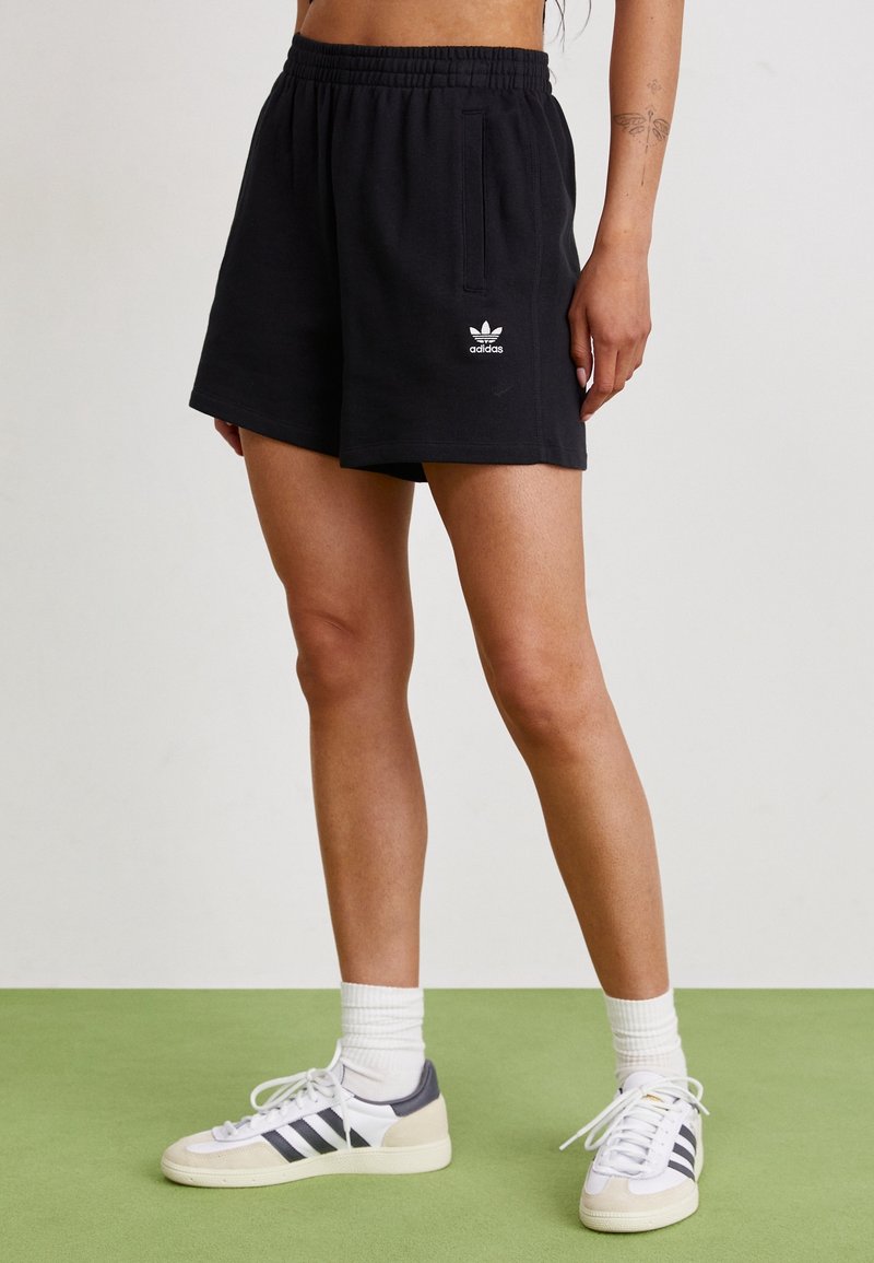 Black cotton shorts with an elastic waistband and side pockets, featuring a white Adidas logo. Paired with white and gray sneakers.