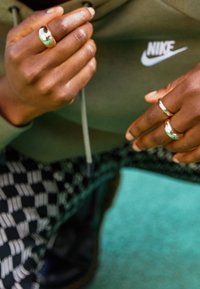 Złote i srebrne metalowe pierścienie na palcach, ubrana w szarą bluzę z kapturem oraz czarne spodnie w wzory. Widoczne są gładkie powierzchnie i refleksyjne akcenty.