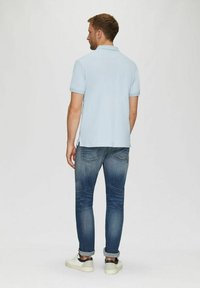 Mann in hellblauem Polohemd, ausgewaschenen blauen Jeans mit umgeschlagenen Säumen und weißen Turnschuhen, der mit dem Rücken zur Kamera vor einem schlichten weißen Hintergrund steht.
