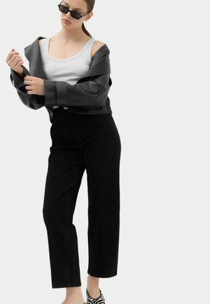 Kvinde iført solbriller, sort kort jakke over grå tanktop, sorte bukser og sko med zebra mønster, poserer foran en ensfarvet baggrund.