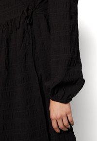 Svart klänning med texturerad tyg, med ett knyt i midjan och volangärmar. Designen inkluderar ett subtilt randigt mönster.