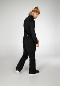 Maglietta nera a maniche lunghe e pantaloni da sci neri con cerniere. Scarponi da sci bianchi. Berretto marrone e bianco a fantasia. Modello in posizione leggermente laterale.