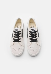 Baskets en daim blanc avec accents noirs, dotées de lacets noirs, de détails perforés au niveau des orteils, et d'un logo Vans sur la languette.