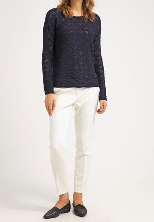 Femme portant un pull texturé bleu marine à manches longues, un pantalon blanc slim fit et des mocassins noirs pointus, debout sur un fond uni.