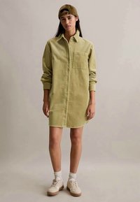 Mulher vestindo um vestido de corduroy verde claro com botões, ténis bege, meias brancas e um boné de basebol castanho ao contrário, de pé contra um fundo simples.