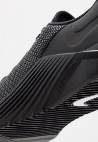 Scarpa atletica nera con tomaia in mesh, suola in gomma, dettagli testurizzati e un logo prominente. Presenta un design aerodinamico e un motivo di presa.