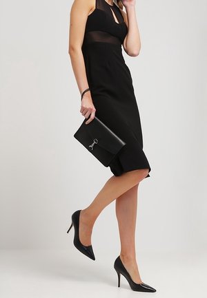 Vestito nero senza maniche con scollatura a halter e parte superiore trasparente. Abbinato a scarpe con tacco alto nere e una pochette nera con chiusura.