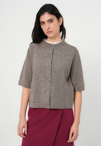 Καφέ ζακέτα με κοντά μανίκια κατασκευασμένη από απαλό ύφασμα, με στρογγυλή λαιμόκοψη και έξι κουμπιά στο μπροστινό μέρος. Φορεμένη πάνω από μια μπορντό φούστα.