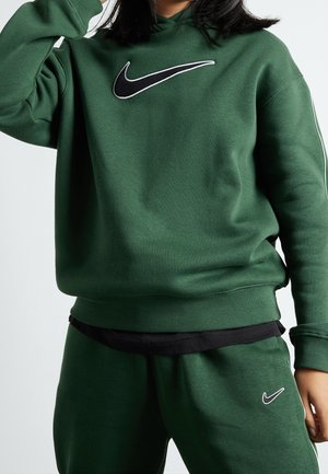 Personne portant un sweat-shirt Nike vert foncé et un pantalon assorti avec des logos Nike noirs et blancs, debout devant un fond clair uni.