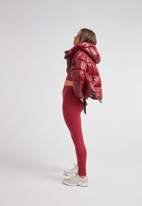 Giacca a vento rossa con cappuccio, texture lucida e zip laterale; abbinata a leggings rossi coordinati. Le scarpe da ginnastica sono beige con dettagli riflettenti.