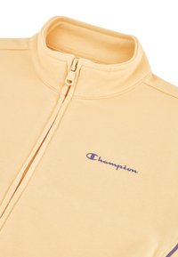 Chaqueta de forro polar amarilla claro con cremallera y cuello alzado, que presenta un logo morado de "Champion" en el lado izquierdo del pecho y un acabado de tejido texturizado.