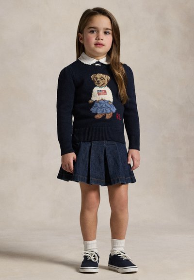 Junges Mädchen steht, trägt einen marineblauen Pullover mit Bärendesign, weißes Hemd mit Kragen, plissierten Jeansrock, weiße Socken und marineblaue Sneakers