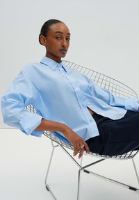 Camicia button-up azzurro chiaro con una vestibilità rilassata, grandi polsini e colletto classico, indossata sopra pantaloni scuri mentre si è seduti su una sedia in filo.