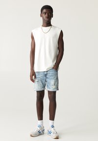 White sleeveless shirt, ripped light blue denim shorts, grey sneakers with blue accents, standing pose, short dark hair, simple gold chain.