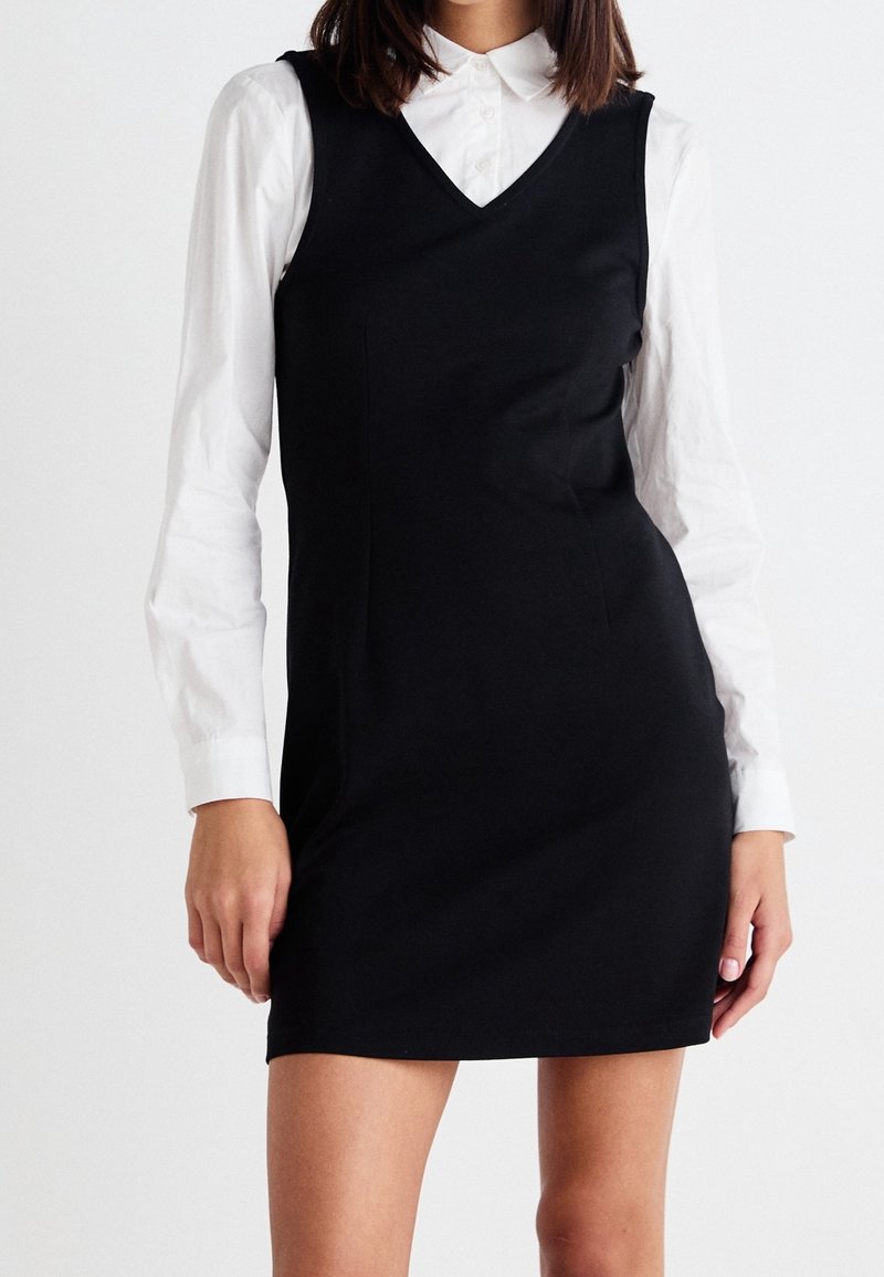 Femme portant une robe noire sans manches col en V par-dessus une chemise blanche à manches longues, debout devant un fond blanc uni.