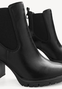 Paire de bottines en cuir noir avec talons épais, panneaux latéraux élastiques et languettes de tirage sur fond blanc.