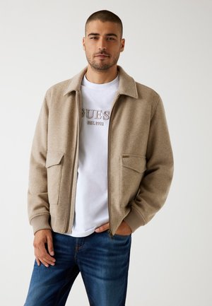 Beige ritsjack met een zachte textuur, twee voorkruiszakken, geribbelde manchetten en een kraag over een wit T-shirt en blauwe spijkerbroek.