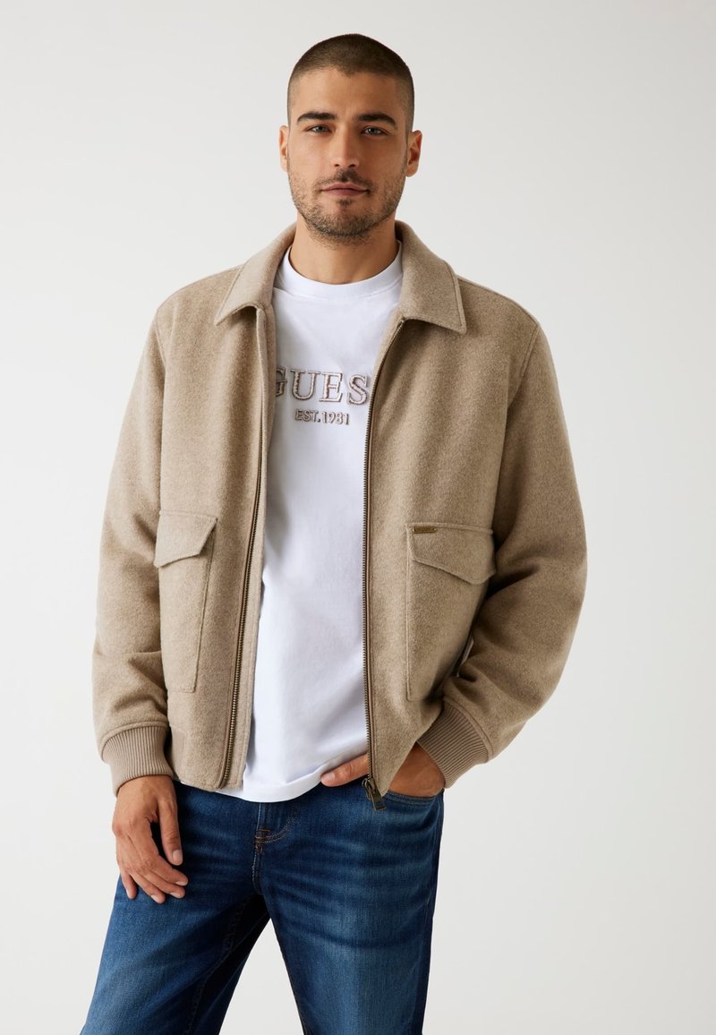 Beige ritsjack met een zachte textuur, twee voorkruiszakken, geribbelde manchetten en een kraag over een wit T-shirt en blauwe spijkerbroek.