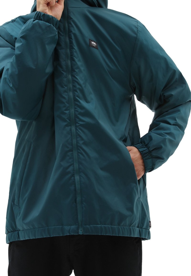 Homme portant une veste zippée bleu sarcelle avec capuche, une main ajustant le col, l’autre main dans la poche, sur fond blanc.