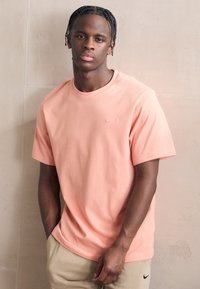 T-shirt Nike rose clair à manches courtes, col rond et logo sur la poitrine ; associé à un pantalon beige ; texture en coton lisse.