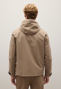 Giacca impermeabile beige con cappuccio, caratterizzata da una texture liscia, cuciture minime e polsini regolabili. Adatta a diverse condizioni atmosferiche all'aperto.