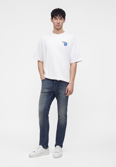 Νέος άνδρας που στέκεται, φοράει λευκό T-shirt με μικρό μπλε λογότυπο, ξεβαμμένα μπλε τζιν και λευκά αθλητικά παπούτσια σε απλό φόντο.