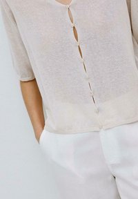 Hellbeige, transparente Bluse mit Dreiviertelärmeln, V-Ausschnitt und einer zentralen Knopfleiste mit kleinen Knöpfen, kombiniert mit weißen Hosen.