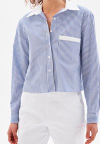 Donna che indossa una camicia a righe verticali blu e bianche con bordo della tasca bianca, abbinata a pantaloni bianchi a vita alta.