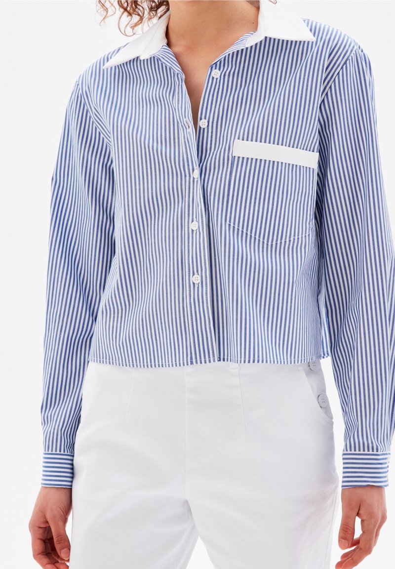 Donna che indossa una camicia a righe verticali blu e bianche con bordo della tasca bianca, abbinata a pantaloni bianchi a vita alta.