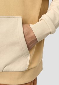 Beige och creamfärgad bomullshoodie med en känguruficka, ribbade muddar och en slät yta som framhäver kontrasterande färgsektioner.