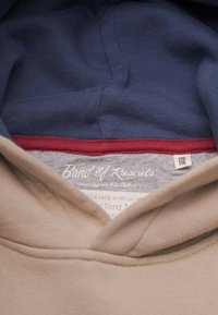 Beige hoodie inredning med strukturerad marinblå foder och röd accent. Grå etikett med texten "Band of Rascals," med storlek och skötselinstruktioner synliga.